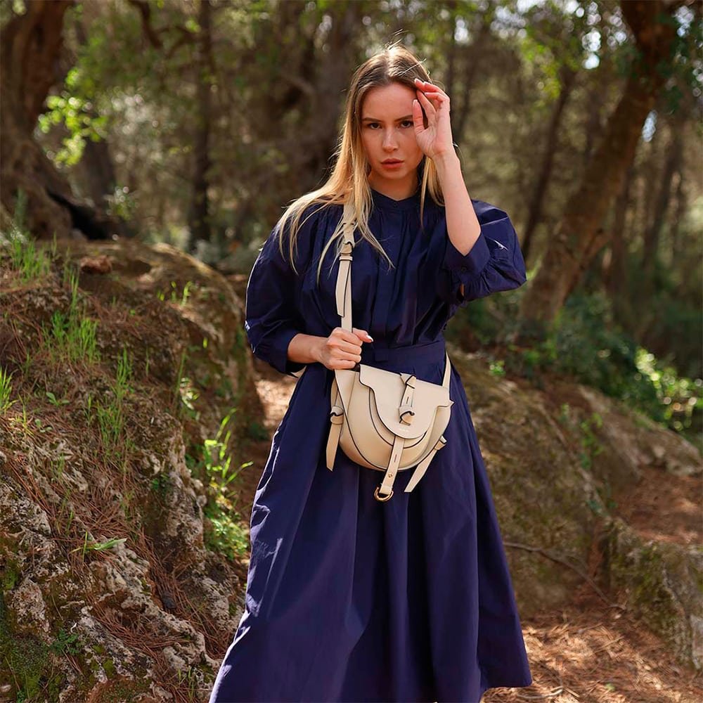 Mujer con vestido azul marino en un entorno natural, luciendo un bolso bandolera Lauburu en color blanco hueso. Diseño sofisticado con detalles en negro, correa ajustable y hebilla dorada. Accesorio elegante y versátil para cualquier ocasión