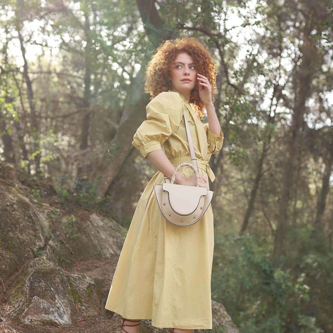 Mujer pelirroja con vestido amarillo y bolso Lauburu blanco en un bosque soleado. Moda sostenible y elegante con accesorios de cuero artesanales