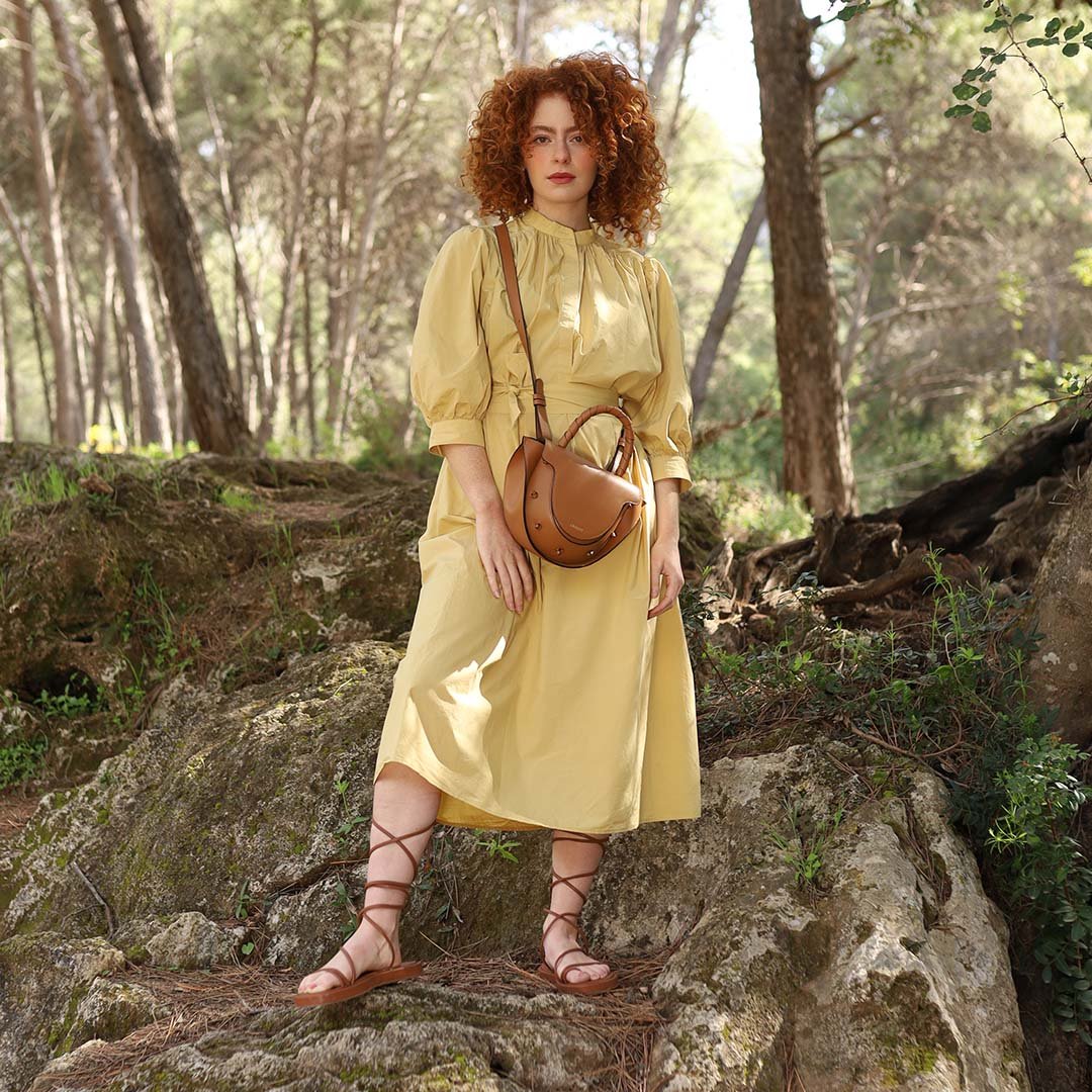 Mujer pelirroja con vestido amarillo y sandalias romanas posando en un entorno natural con un bolso Lauburu en color camel con asa trenzada. Estilo bohemio y sofisticado en un escenario forestal