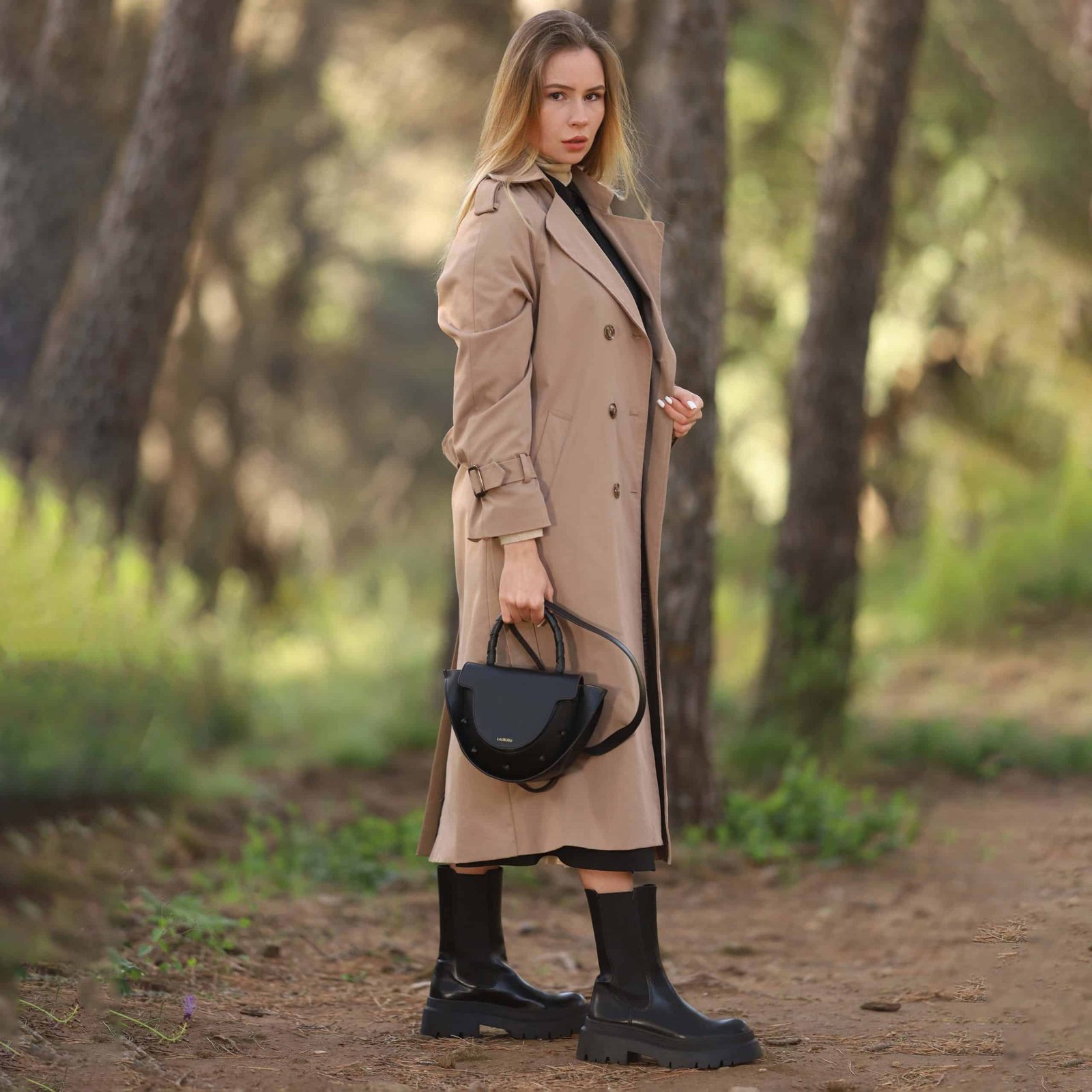 Mujer rubia con abrigo trench beige, vestido negro y botas altas de suela gruesa, sosteniendo un bolso Lauburu negro con asa trenzada en un entorno natural con árboles y luz tenue. Look elegante y sofisticado con inspiración otoñal