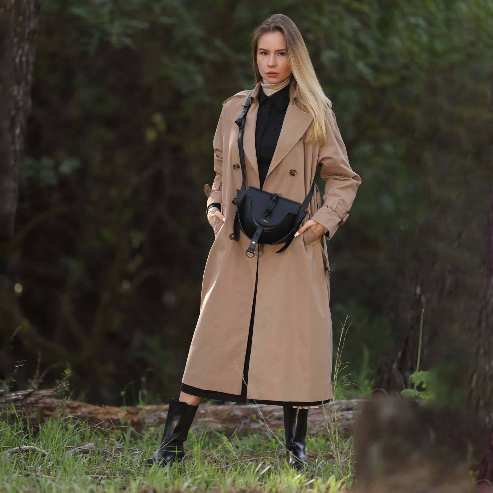 Mujer rubia con abrigo trench beige y vestido negro, combinados con botas altas de cuero y un bolso Lauburu negro cruzado. Look elegante y sofisticado en un entorno natural con fondo boscoso