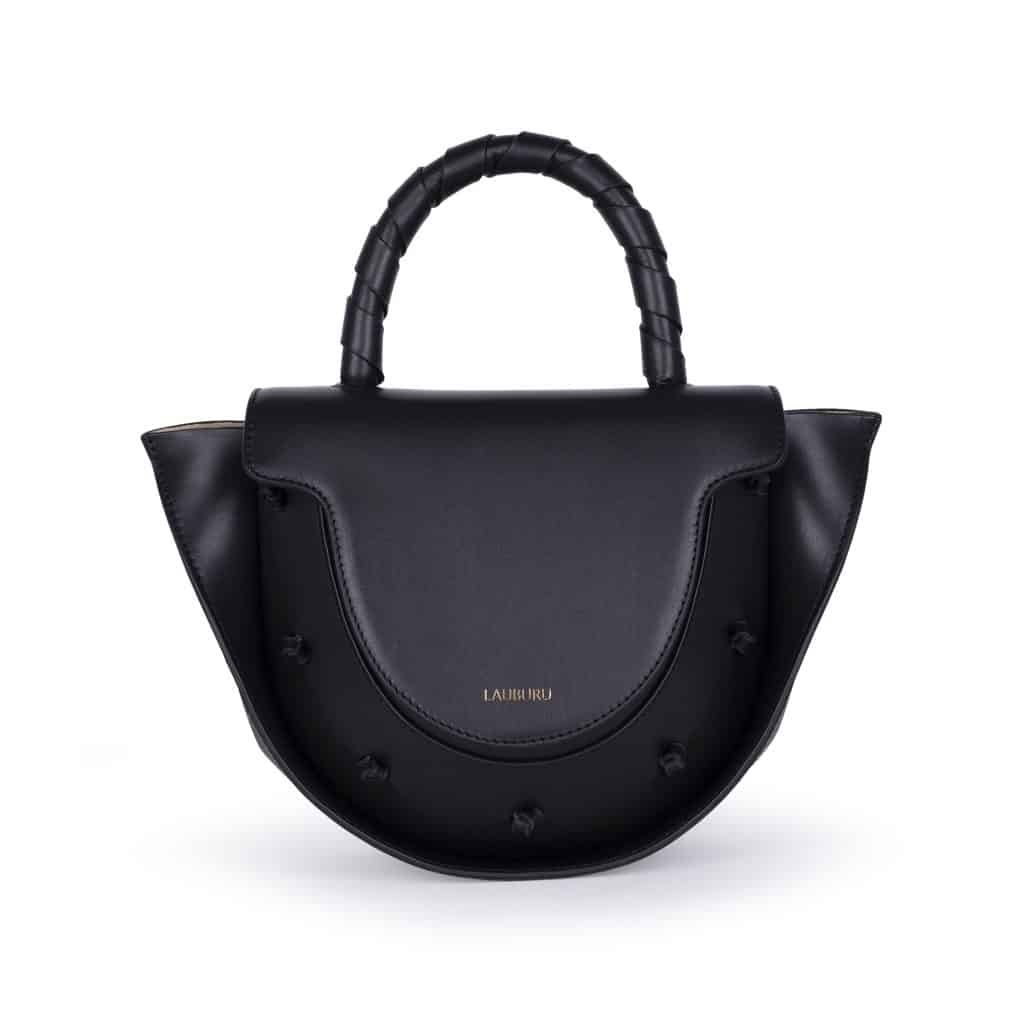 Bolso Lauburu en piel negra con asa trenzada y detalles de nudos en la solapa. Diseño estructurado y sofisticado con logotipo dorado grabado y bordes en contraste. Perfecto para un look elegante y versátil