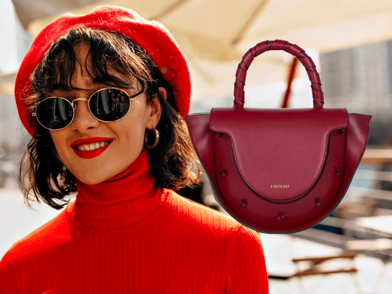 Mujer con boina roja, gafas de sol redondas y suéter de cuello alto rojo, sonriendo en un ambiente urbano. A su lado, bolso Lauburu burdeos con asa trenzada y detalles metálicos