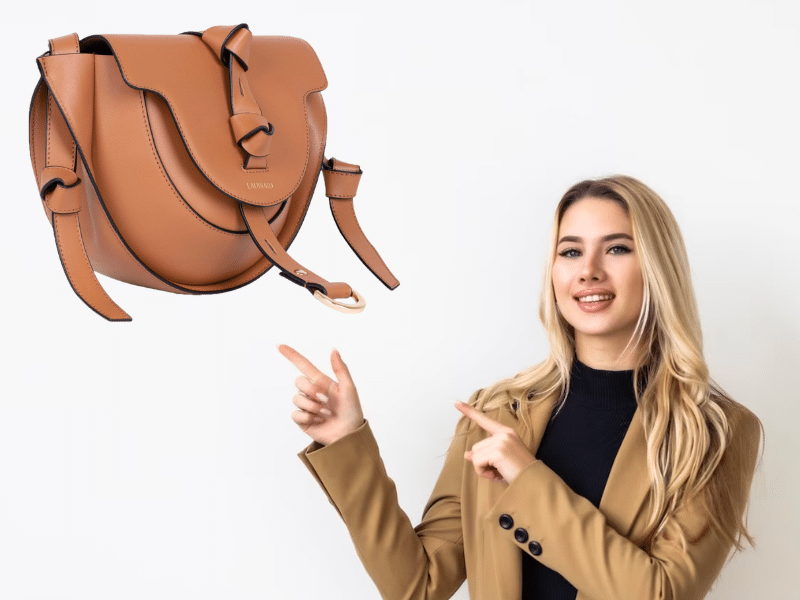 Mujer rubia con blazer camel y suéter negro señalando un bolso Lauburu en tono cuero flotante sobre fondo blanco. Diseño elegante con detalles de nudos y herrajes dorados
