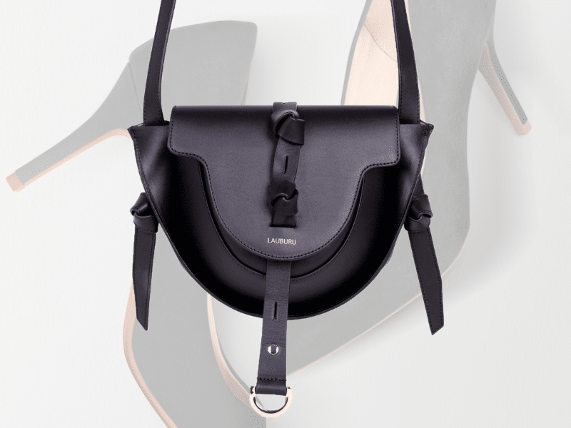 Bolso negro Lauburu con diseño de nudos y detalles dorados, flotando sobre un fondo con tacones difuminados en tonos beige y negro. Estilo sofisticado y moderno en accesorios de moda