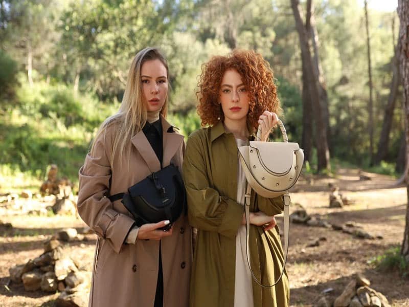 Dos mujeres en un bosque con abrigos elegantes y bolsos Lauburu de cuero. Una lleva un bolso negro cruzado, mientras que la otra sostiene un bolso blanco con asa trenzada, destacando la artesanía y el diseño sofisticado