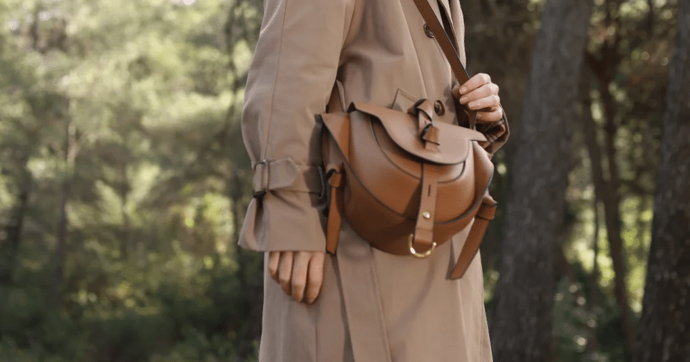 Persona con gabardina beige en un bosque, luciendo un bolso de cuero marrón Lauburu con detalles de nudos y hebilla dorada. Elegancia y diseño artesanal