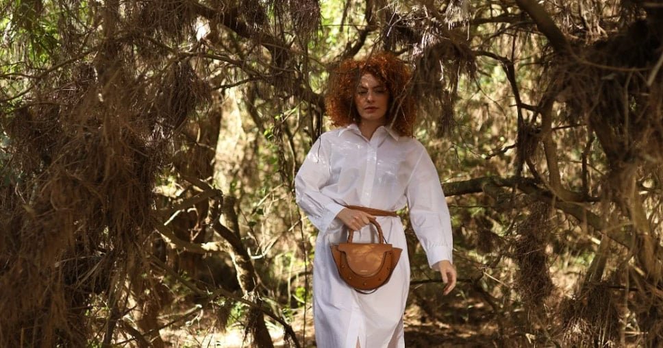 Mujer con vestido blanco camina por un bosque sosteniendo un bolso de cuero marrón de Lauburu con asa trenzada. Diseño sofisticado con detalles artesanales