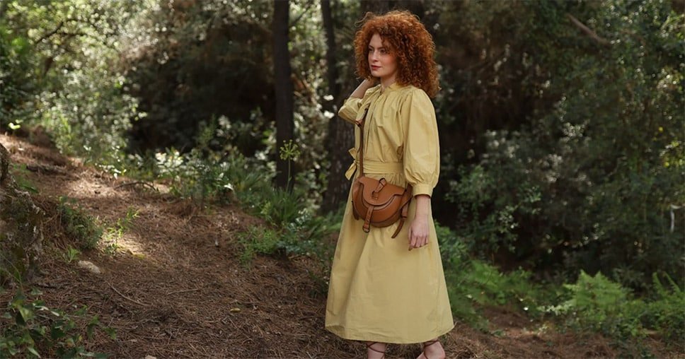 Mujer pelirroja con vestido amarillo en un bosque, llevando un bolso de cuero marrón de Lauburu con diseño artesanal y correa ajustable