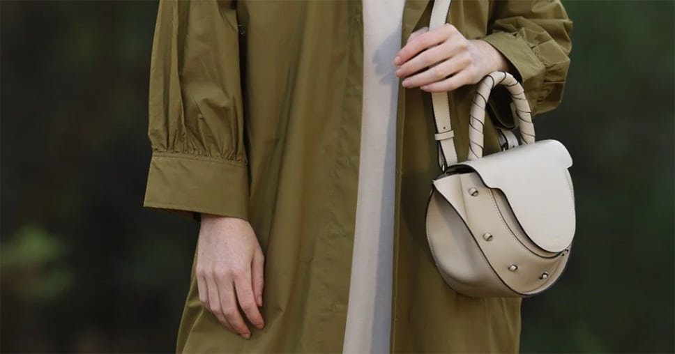 Primer plano de una mujer con abrigo verde sosteniendo un bolso de cuero beige de Lauburu, con asa trenzada y detalles metálicos, en un entorno natural