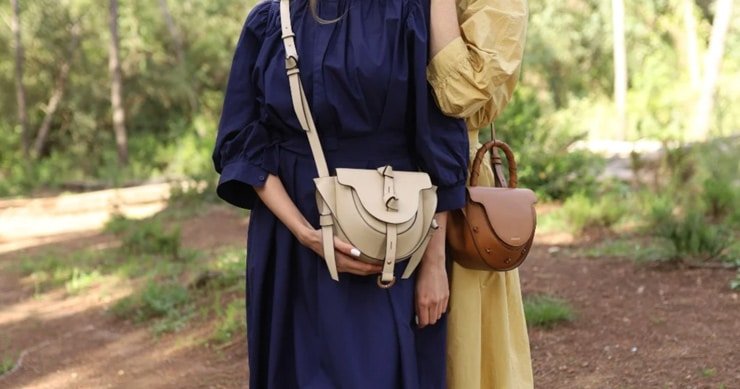 Dos mujeres con vestidos elegantes en tonos azul y amarillo, luciendo bolsos de cuero Lauburu en color beige y marrón, en un entorno natural y luminoso