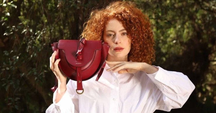Mujer de cabello rizado pelirrojo con camisa blanca sosteniendo un bolso de cuero rojo de la marca Lauburu, con diseño de solapa y detalles de nudos, en un entorno natural con luz cálida