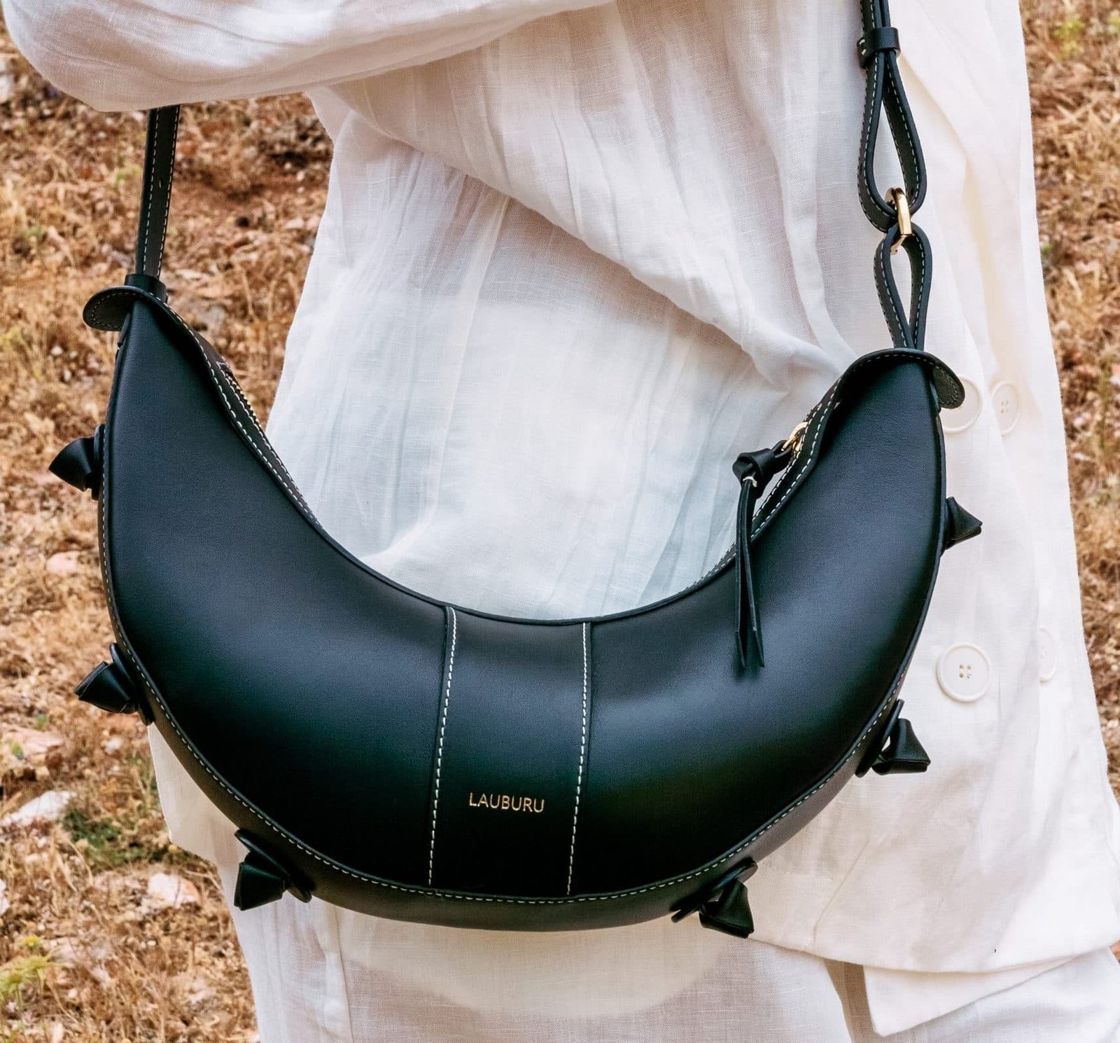 Bolso negro de diseño curvado Lauburu con costuras en contraste y detalles de nudos decorativos, llevado por una persona vestida con un conjunto blanco de lino. Fondo natural con tierra y vegetación seca
