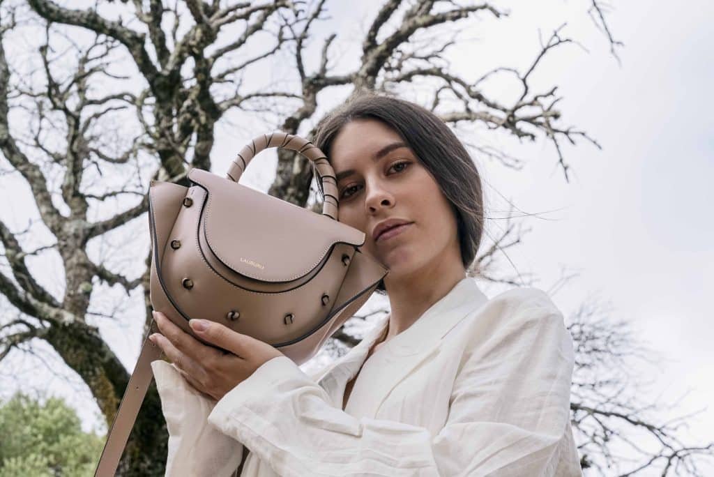 Mujer con blusa blanca sosteniendo un bolso de piel taupe Lauburu con asa trenzada y detalles anudados, en un entorno natural con árboles secos de fondo. Diseño elegante y sofisticado, ideal para un estilo atemporal.