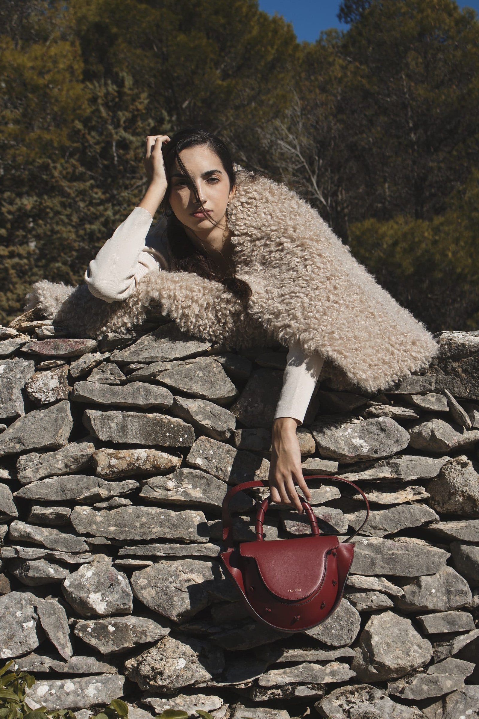 Bolso de piel burdeos Lauburu con asa trenzada y detalles anudados, sostenido por una mujer con abrigo de borrego sobre un muro de piedra en un paisaje natural. Diseño elegante y sofisticado para un estilo exclusivo.