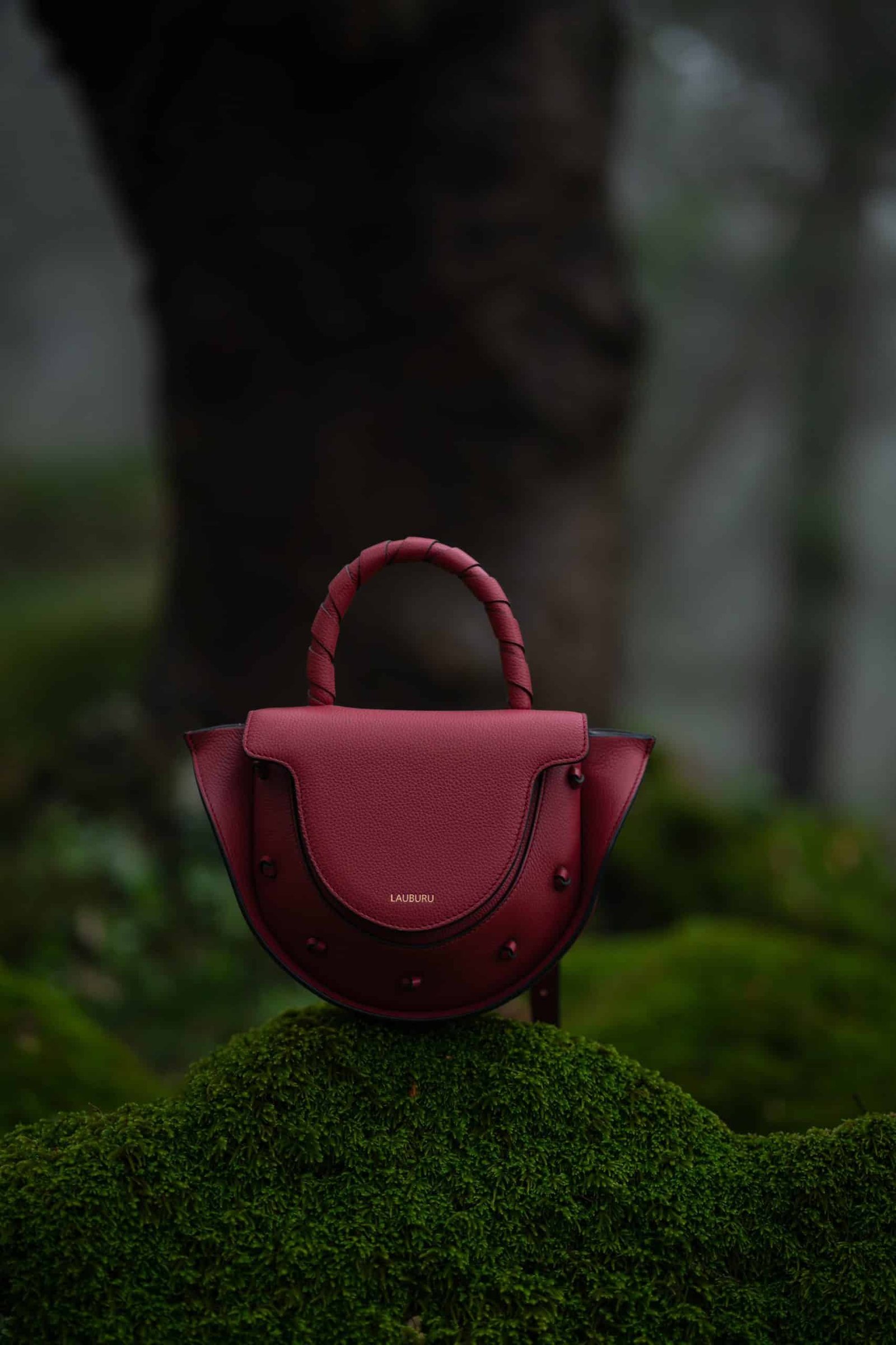 Bolso de piel burdeos Lauburu con asa trenzada y solapa estructurada, posado sobre musgo en un entorno natural. Diseño exclusivo con detalles anudados y cierre magnético, perfecto para un estilo elegante y sofisticado.