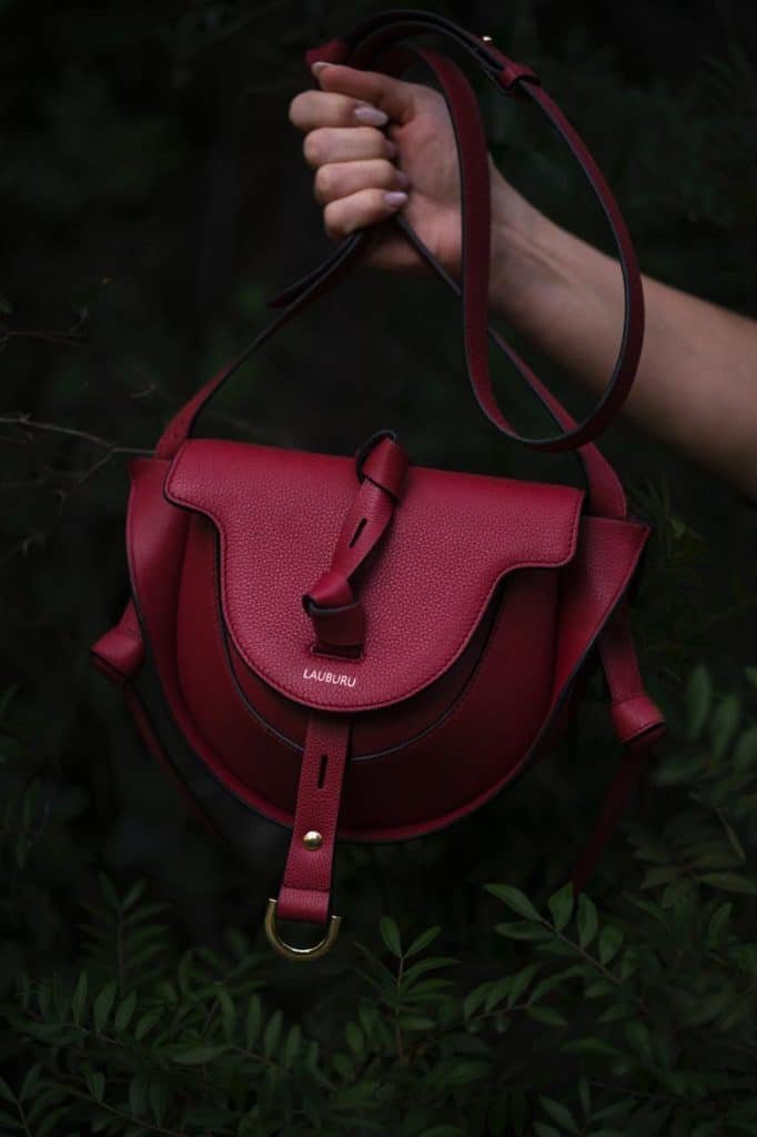 Bolso de piel burdeos Lauburu con solapa estructurada y cierre de hebilla dorada. Diseño elegante con correa ajustable, ideal para un look sofisticado y versátil, capturado en un entorno natural.