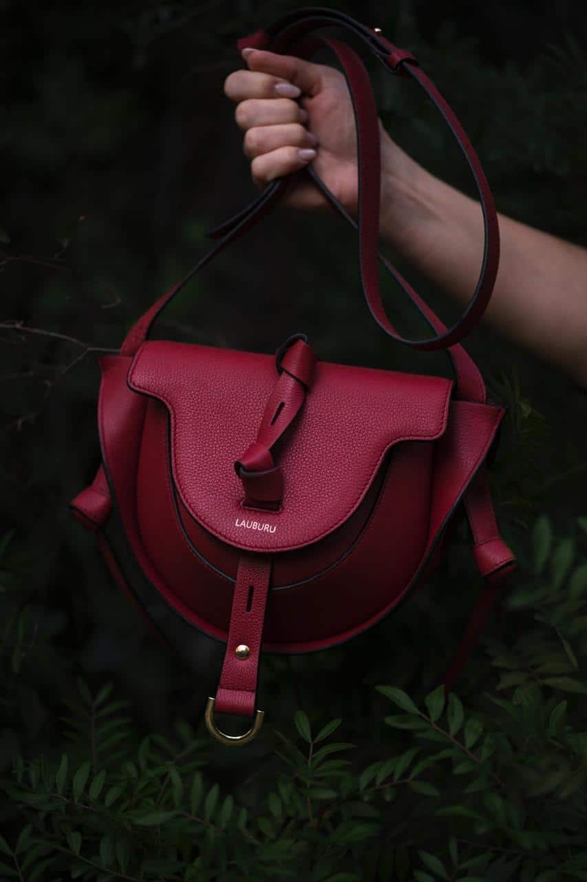 Bolso de piel burdeos Lauburu con solapa estructurada y cierre de hebilla dorada. Diseño elegante con correa ajustable, ideal para un look sofisticado y versátil, capturado en un entorno natural.